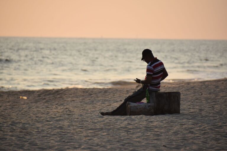 Bas-Congo Et Virunga A man sits on a log at a beach, facing the sea, wearing a striped shirt and a cap, looking at a cell phone in his hand.